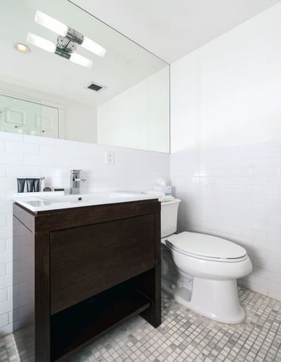 East Village Hotel room with bathroom with tiled walls, dark wooden vanity, white toilet, and wall-mounted mirror.