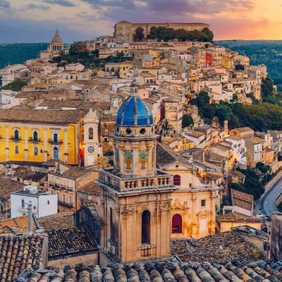 Luftaufnahme der historischen Stadt Matera, Italien, mit blauer Kuppel und gelbem Gebäude bei Sonnenuntergang.