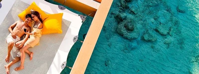A couple relaxing on a sunlit white hammock above clear turquoise water at Novotel Sydney
