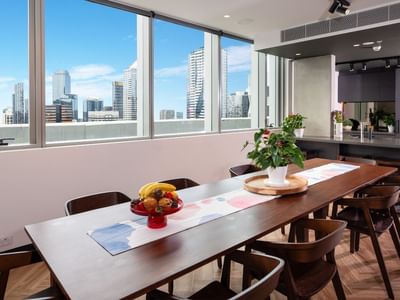 A lounge and dining area with a long table, chairs, fruits, and flowers, with a city skyline view.