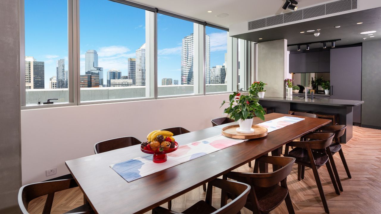 A lounge and dining area with a long table, chairs, fruits, and flowers, with a city skyline view.