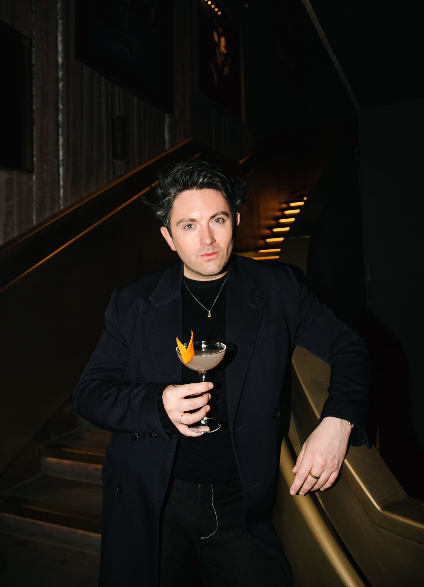 A man with a black coat & a cocktail in a model pose at The Londoner