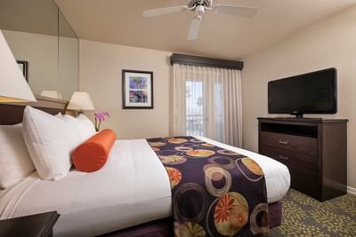 Guest room with white bedding, colorful bed runner, ceiling fan, and flat-screen TV at London Bridge Resort