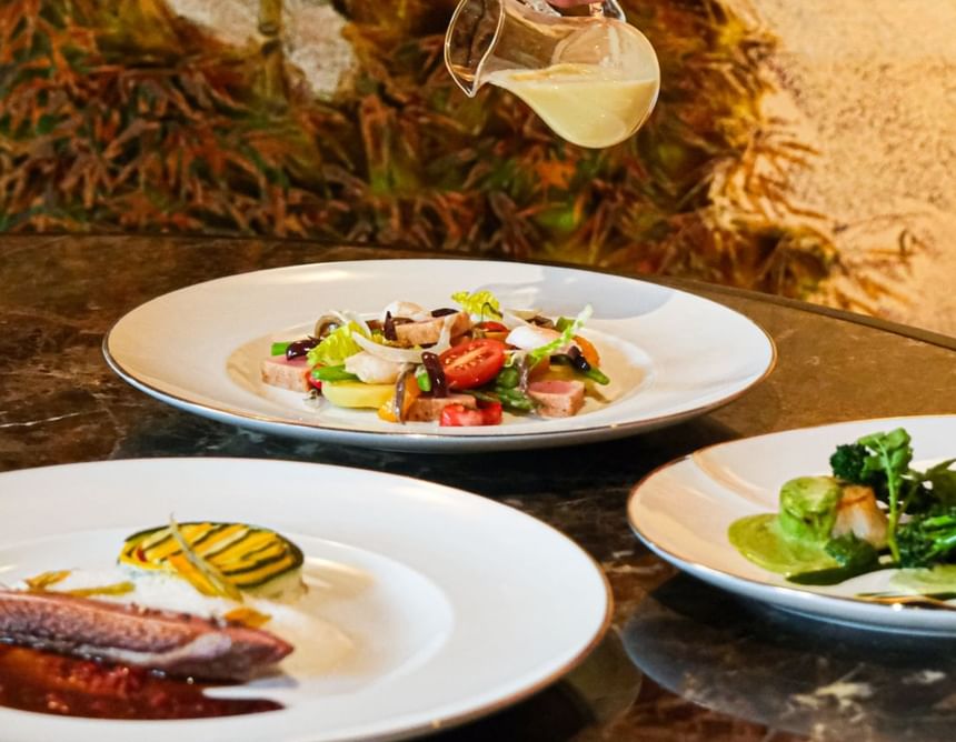 Hand drizzling dressing over a colorful salad on a table with two elegant dishes in Square One at Park Hyatt Saigon