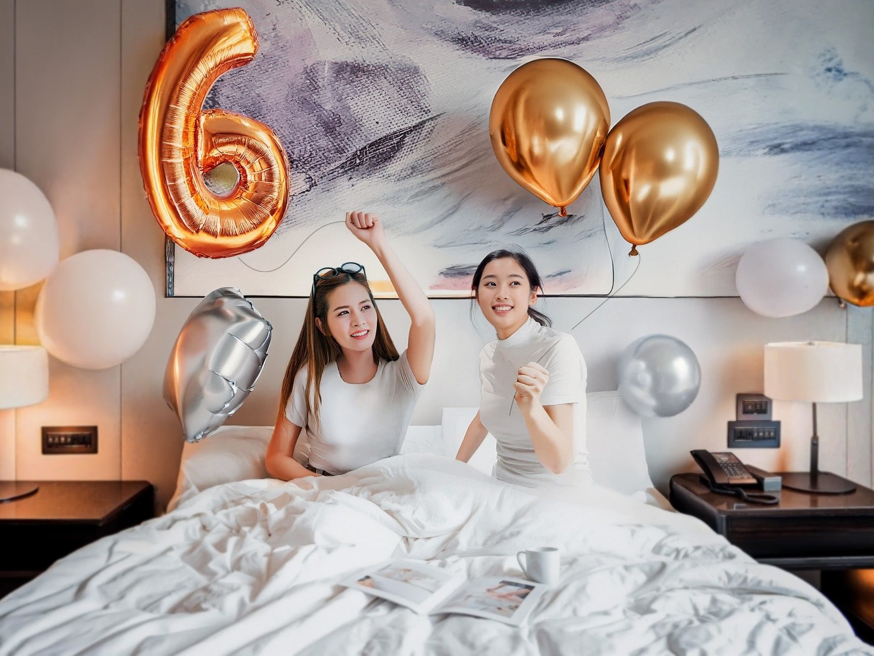 Two women celebrating with balloons and a number 6 on a bed for the 6th Anniversary Celebration.