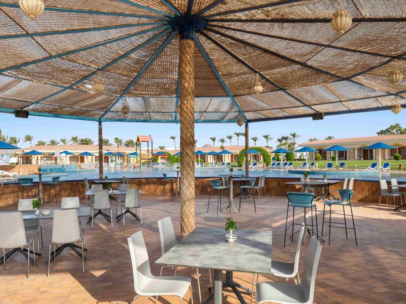 Patio with tables and chairs under a thatched roof, overlooking a pool and resort buildings.