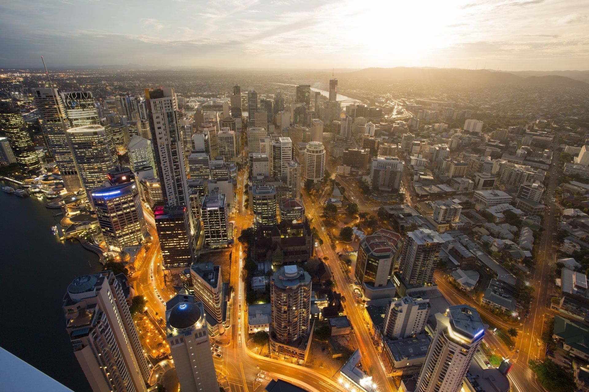 Aerial view of the Brisbane city skyline at dusk, showing illuminated streets and a bright sunset near The Sebel Brisbane