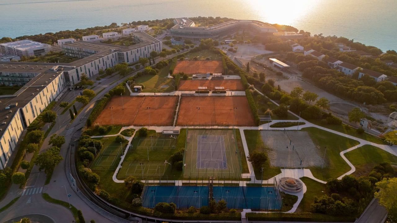 Vista aerea di un resort con campi da tennis e piscina, circondato da edifici e vegetazione.
