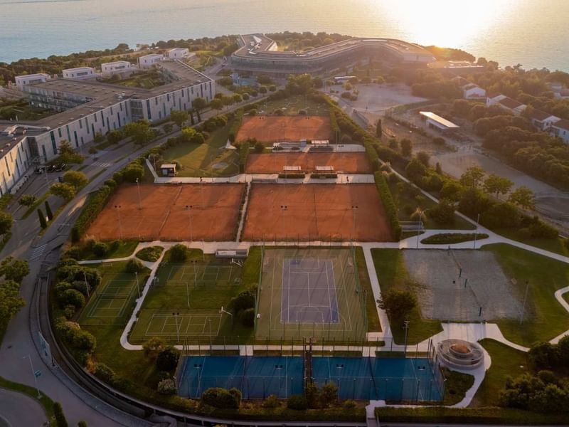 Luftaufnahme eines Tennisclubs mit mehreren Plätzen, Gebäuden und dem Meer im Hintergrund.