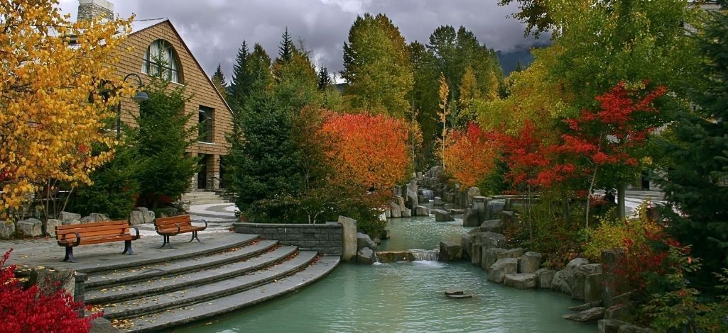 Picturesque pond with autumn-colored trees and rustic architecture at Whistler’s cultural area.
