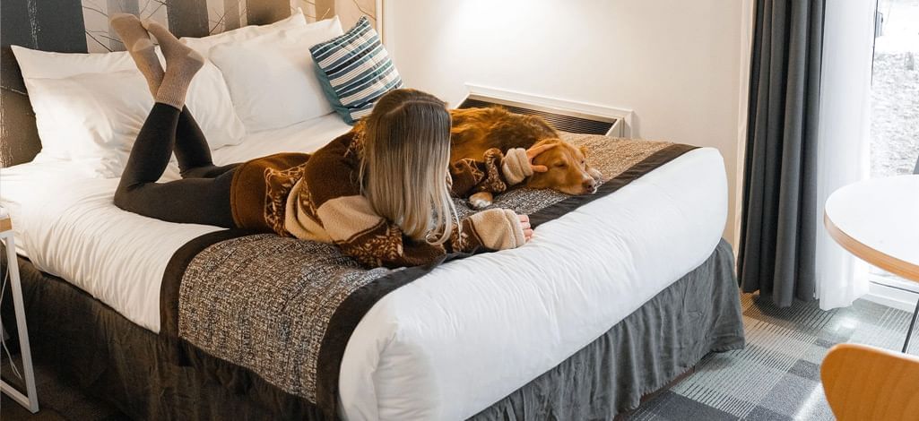 A woman lying on a cozy hotel bed alongside her dog, highlighting the pet-friendly accommodations at Coast Hotels.