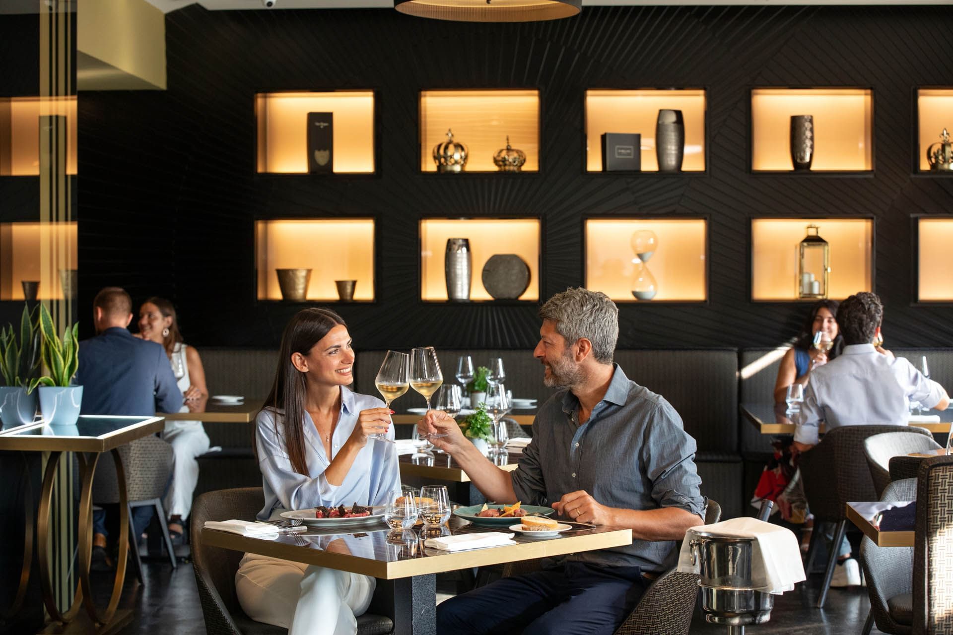 Couple cheers with glasses of wine in a restaurant, surrounded by elegant decor and soft ambient lighting at The Glam Hotel