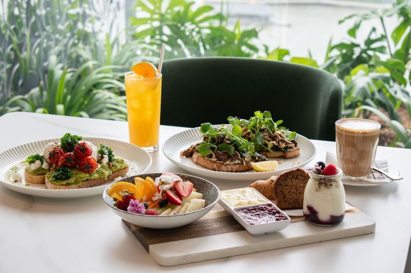 Brunch spread with avocado toast, fresh fruit, and orange drink set against lush greenery at Emporium Hotel South Bank