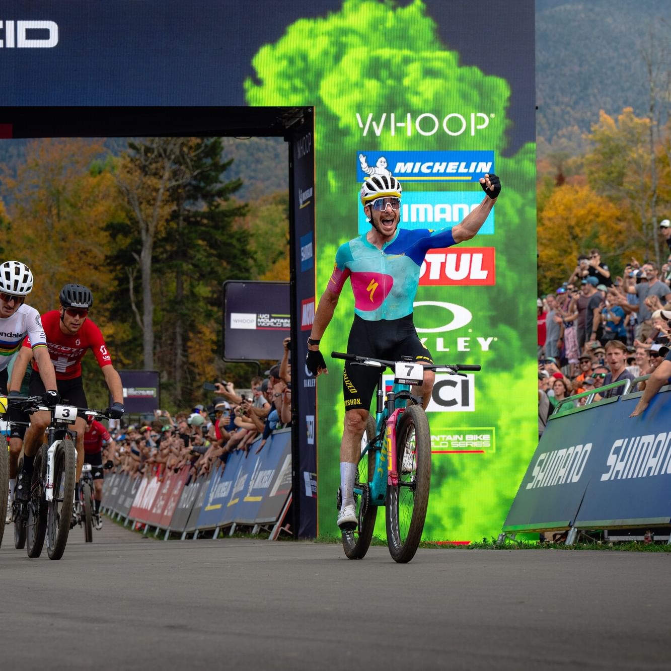 Mountain biker in blue jersey celebrates victory crossing finish line ahead of competitors at WHOOP UCI Mountain Bike World Series