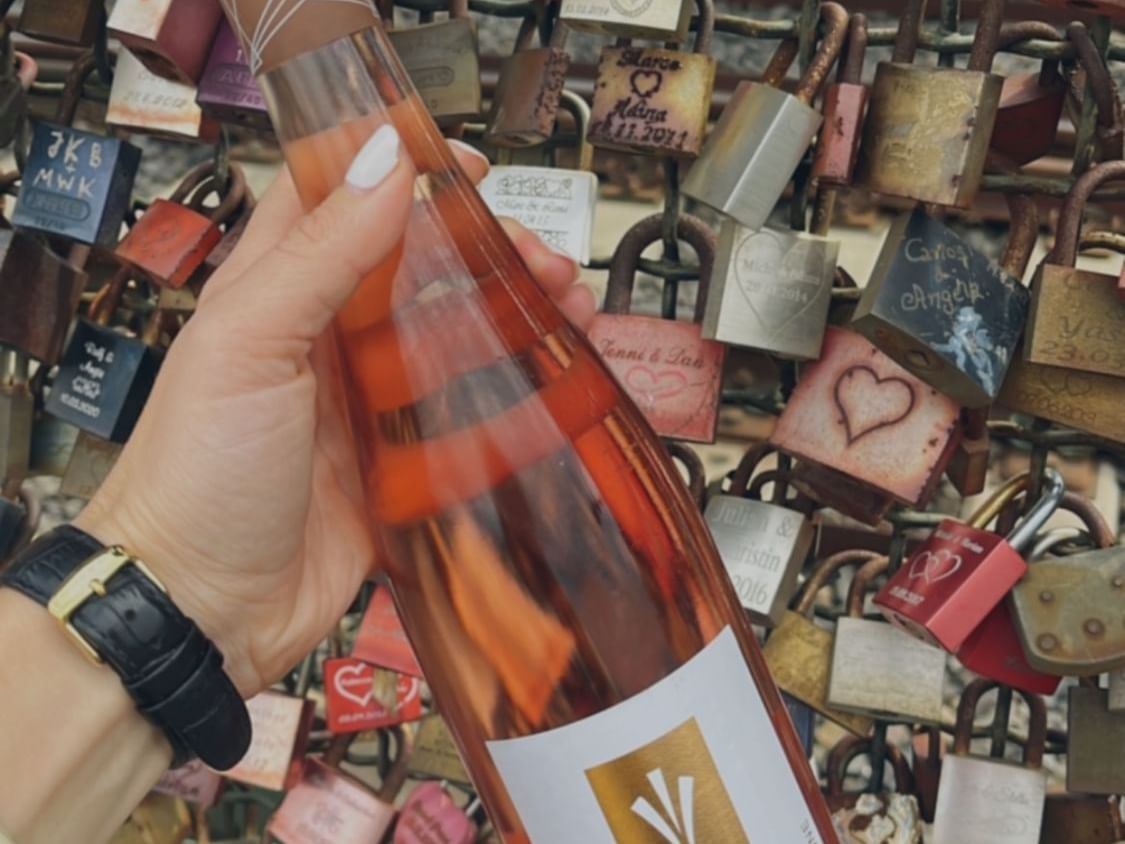 Person holding a bottle of rosé wine in front of a fence with love locks near Classic Hotel Harmonie