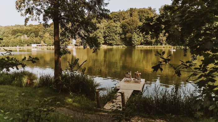 Zwei Personen sitzen auf Liegen an einem Steg neben einem See, umgeben von Bäumen und Gras.