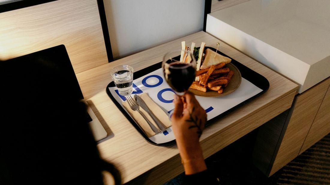 Person with tattooed arm holding wine glass over tray with food and drink on a desk.