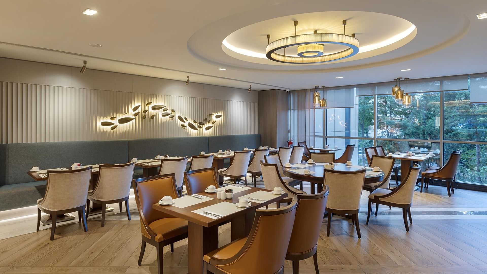 An elegant dining room with wooden tables, cushioned chairs, a decorative ceiling light, and large glass windows in Alesta Restaurant at Titanic Port Bakırköy