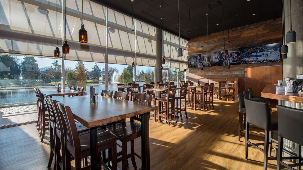 Dining area with wooden tables and chairs, large windows, and a view of the pond at Coast Chilliwack Hotel by APA.