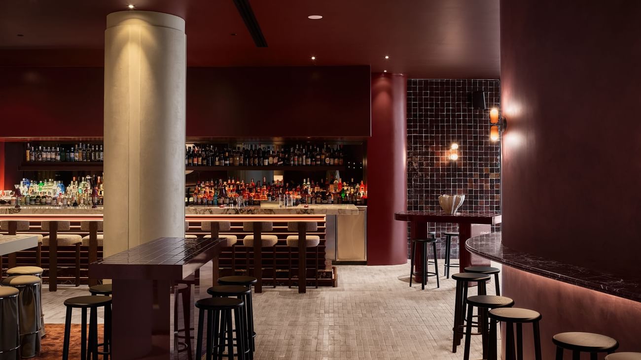 Interior of a bar with wooden tables, stools, and shelves stocked with liquor bottles, with tiled walls and a column.