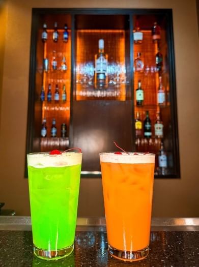 Bright green and bright orange cocktails on a counter in front of a backlit bar at Zebra's Bar and Grill at Rosen Inn at Pointe Orlando.