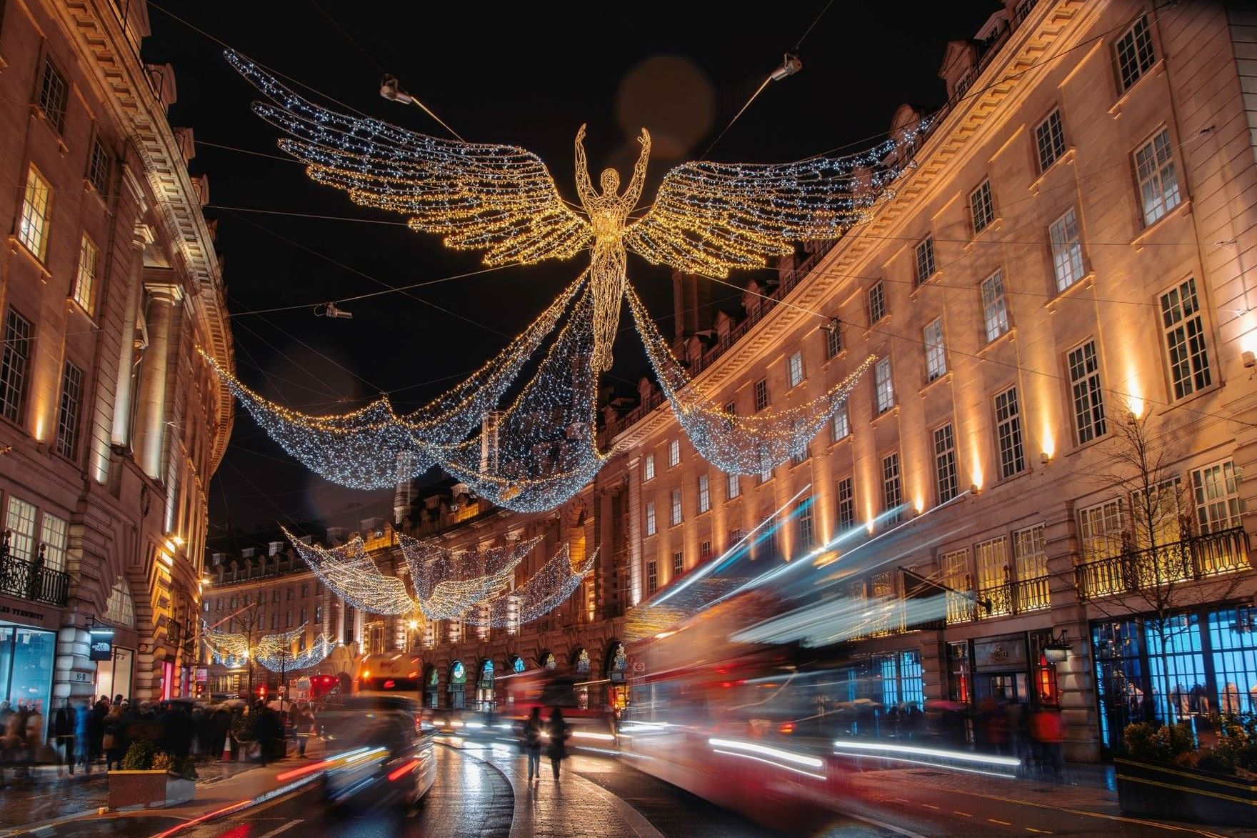 Illuminated angel decorations span across a bustling London street at night with historic buildings
