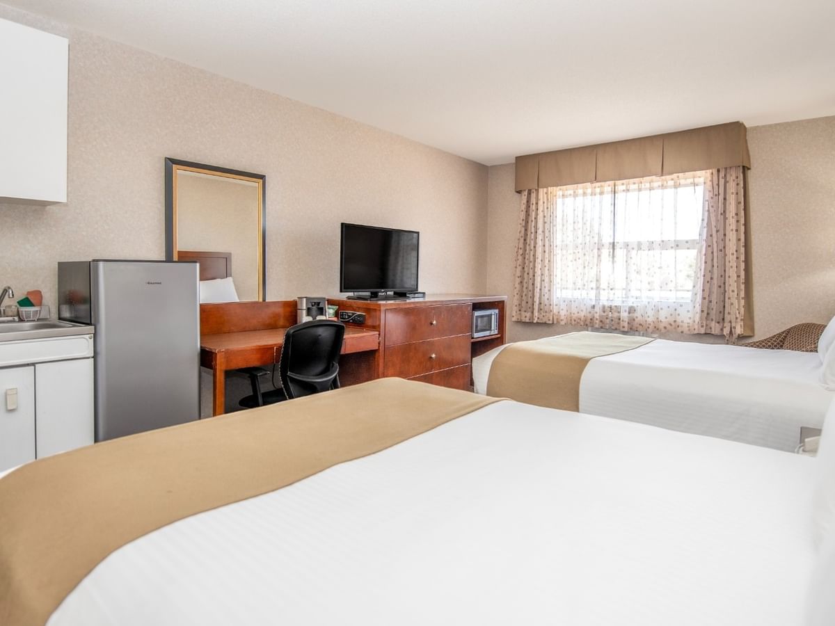 A hotel room with two beds, a desk, a fridge, a sink, a mirror, and a window.