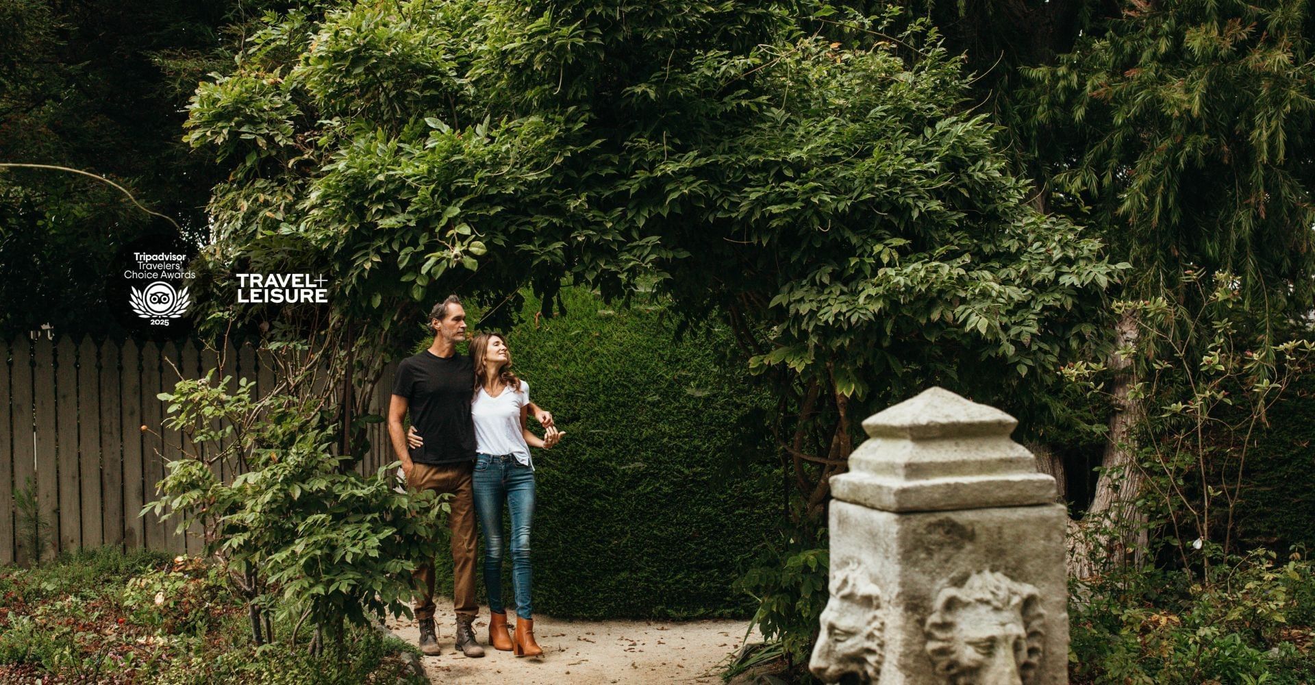 Couple stands smiling in lush garden, framed by greenery and a rustic stone pillar.