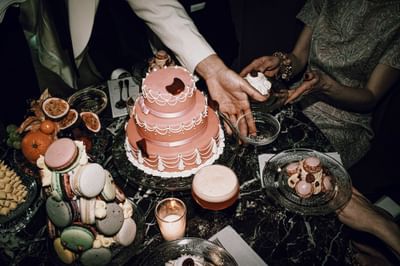 Hand reaching for a pink tiered cake on a dessert table with macarons, fruit, and sparkling drinks at Hotel Motto Vienna