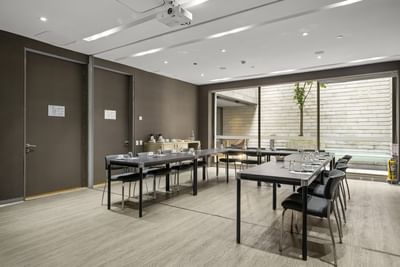 Sala de reuniones con mesas largas, sillas, proyector y ventanas grandes que dan a un patio.