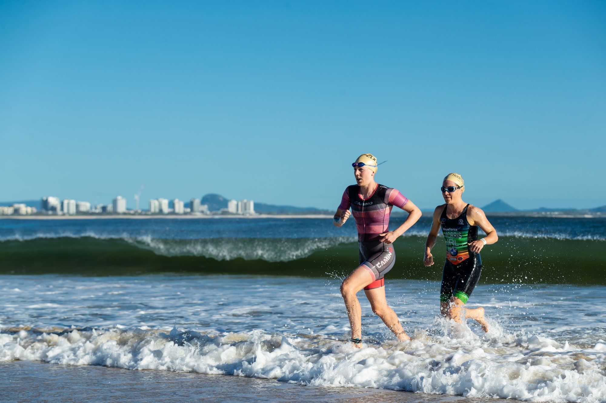 Two athletes in swimsuits and swim caps run through surf near Novotel Sunshine Coast Resort