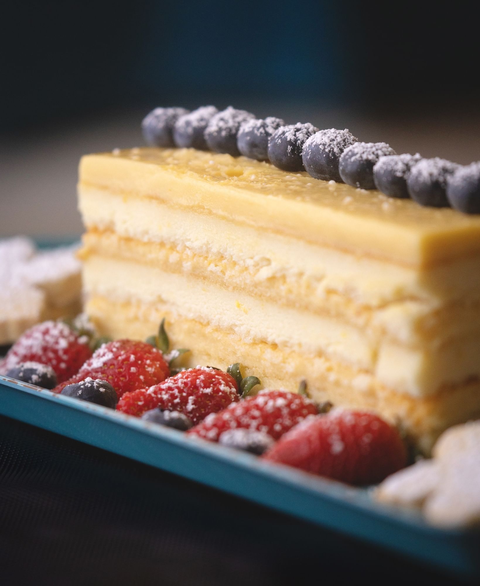 A plated rectangular slice of lemon cake with bluberries lined along the top and strawberries on the side