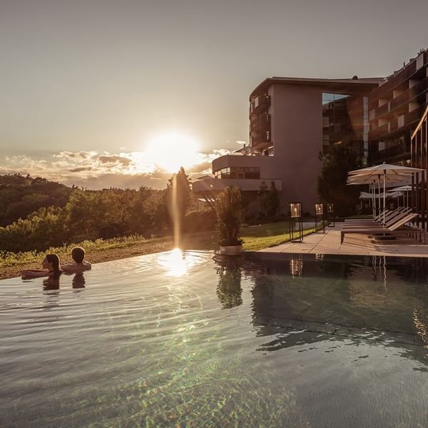 Zwei Personen entspannen im Infinity-Pool bei Sonnenuntergang, umgeben von moderner Architektur und natürlicher Landschaft.