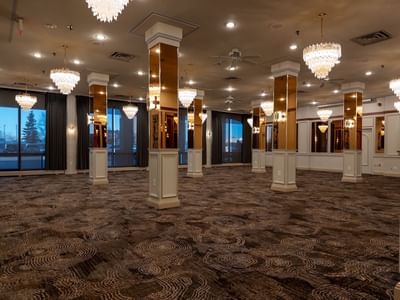 A large, banquet hall at Carriage House with patterned carpet, tall pillars, large windows, and chandeliers.