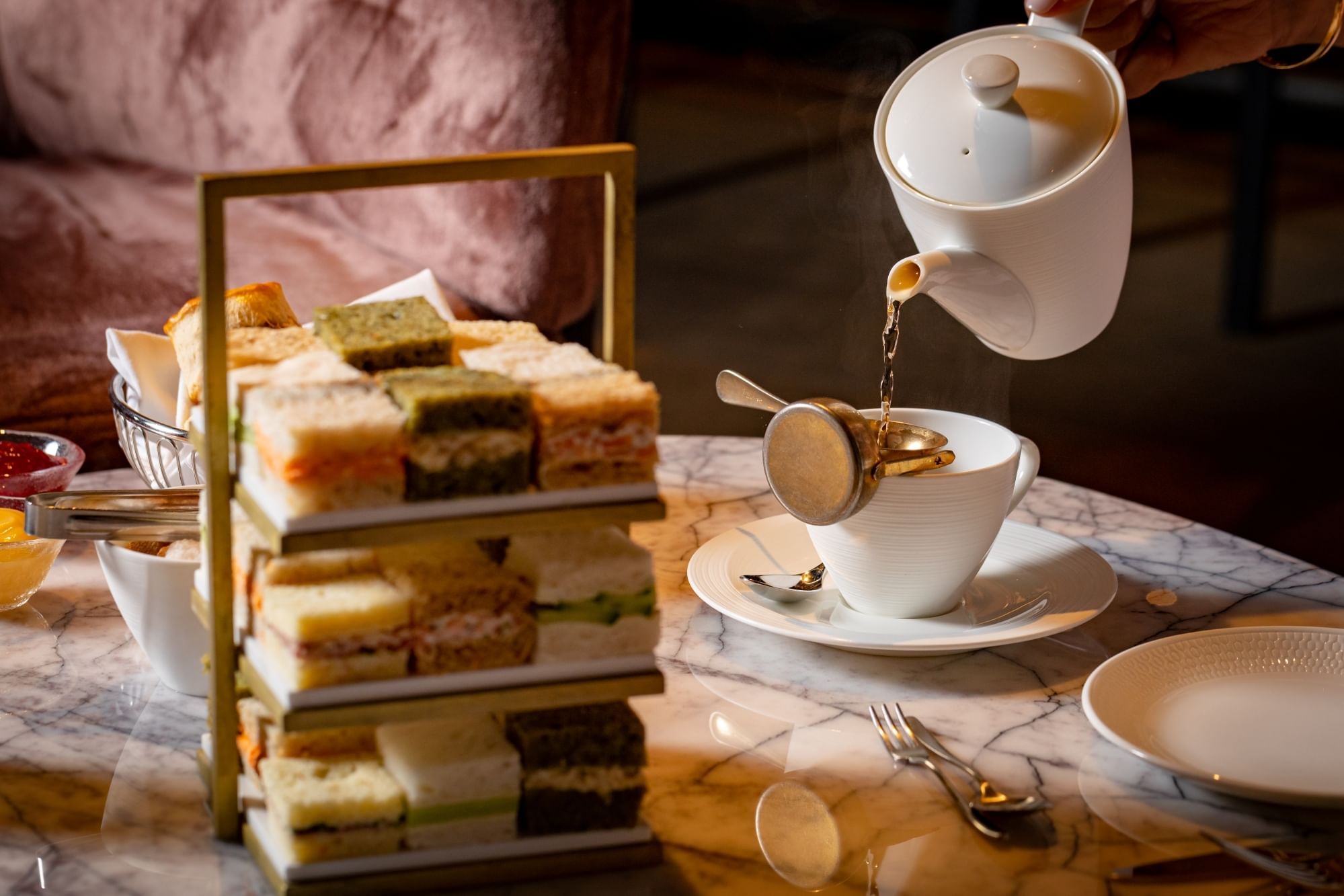 Pouring tea into a cup on the table with pastries and sandwiches at The Londoner Hotel