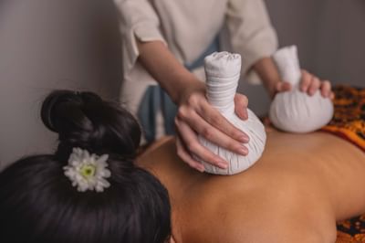 Therapist using white herbal compresses on a guest's back by a floral sarong in LOM Talay Spa at Amora Beach Resort Phuket