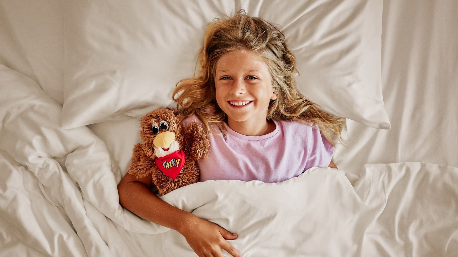 Mädchen liegt im Bett mit Kuscheltier und lacht mit offenem Mund.