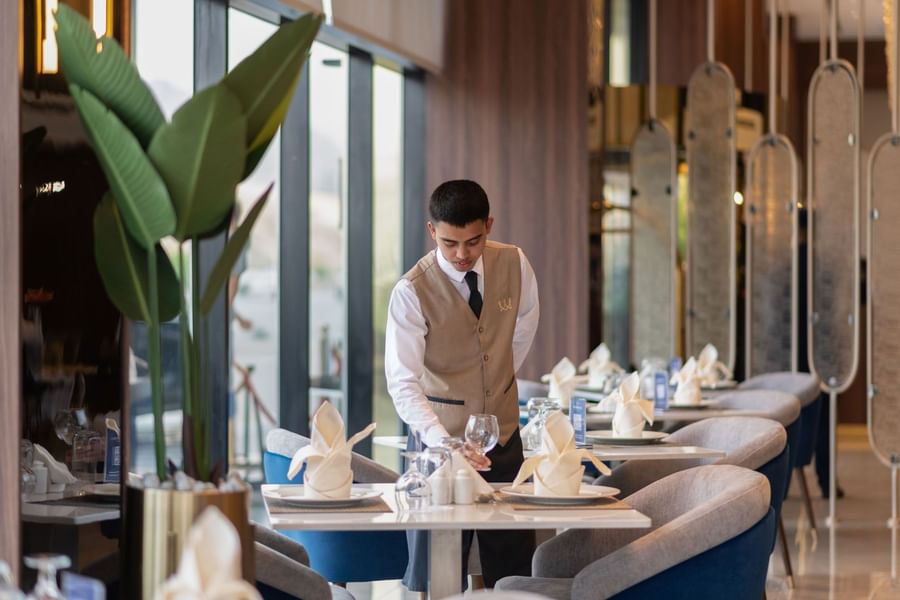 Waiter by a table with blue chairs under wooden wall panels near a plant in Rose Restaurant at Warwick Al Taif Hotel