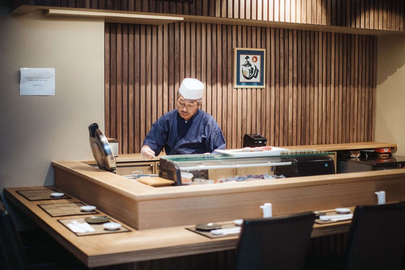 A sushi chef stands behind a wooden sushi bar with a glass display case and several place settings.