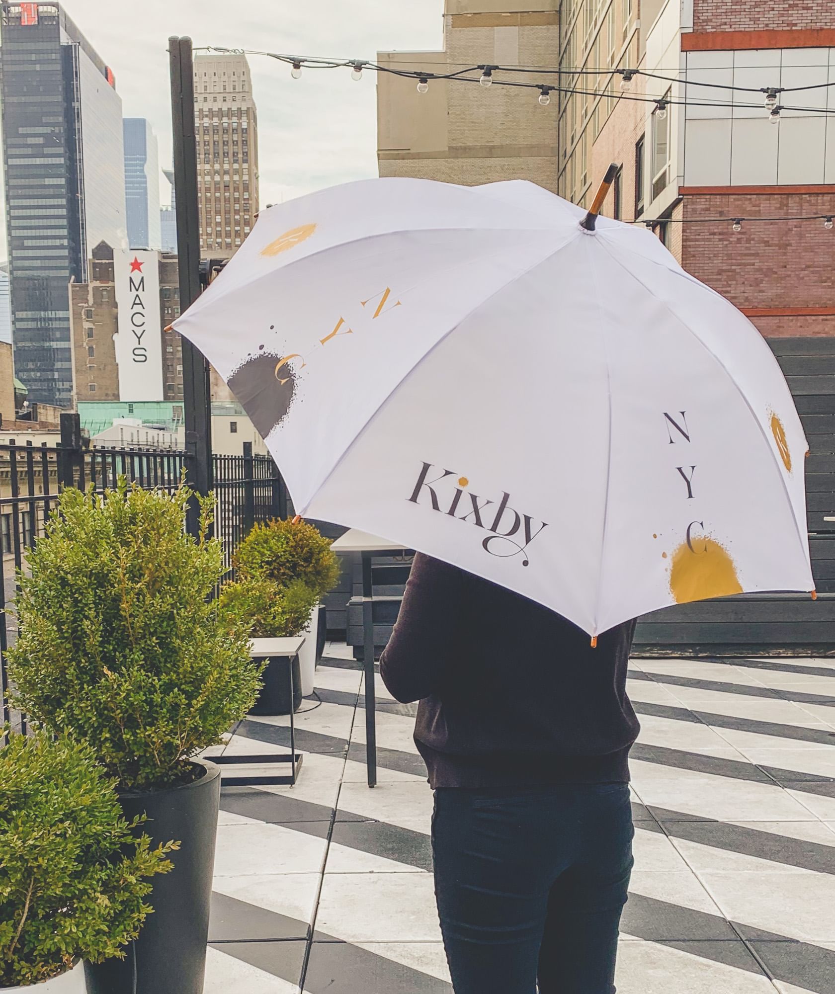 A person holding a white umbrella with the word 'Kixby' on it, standing on a rooftop patio with a city skyline view.