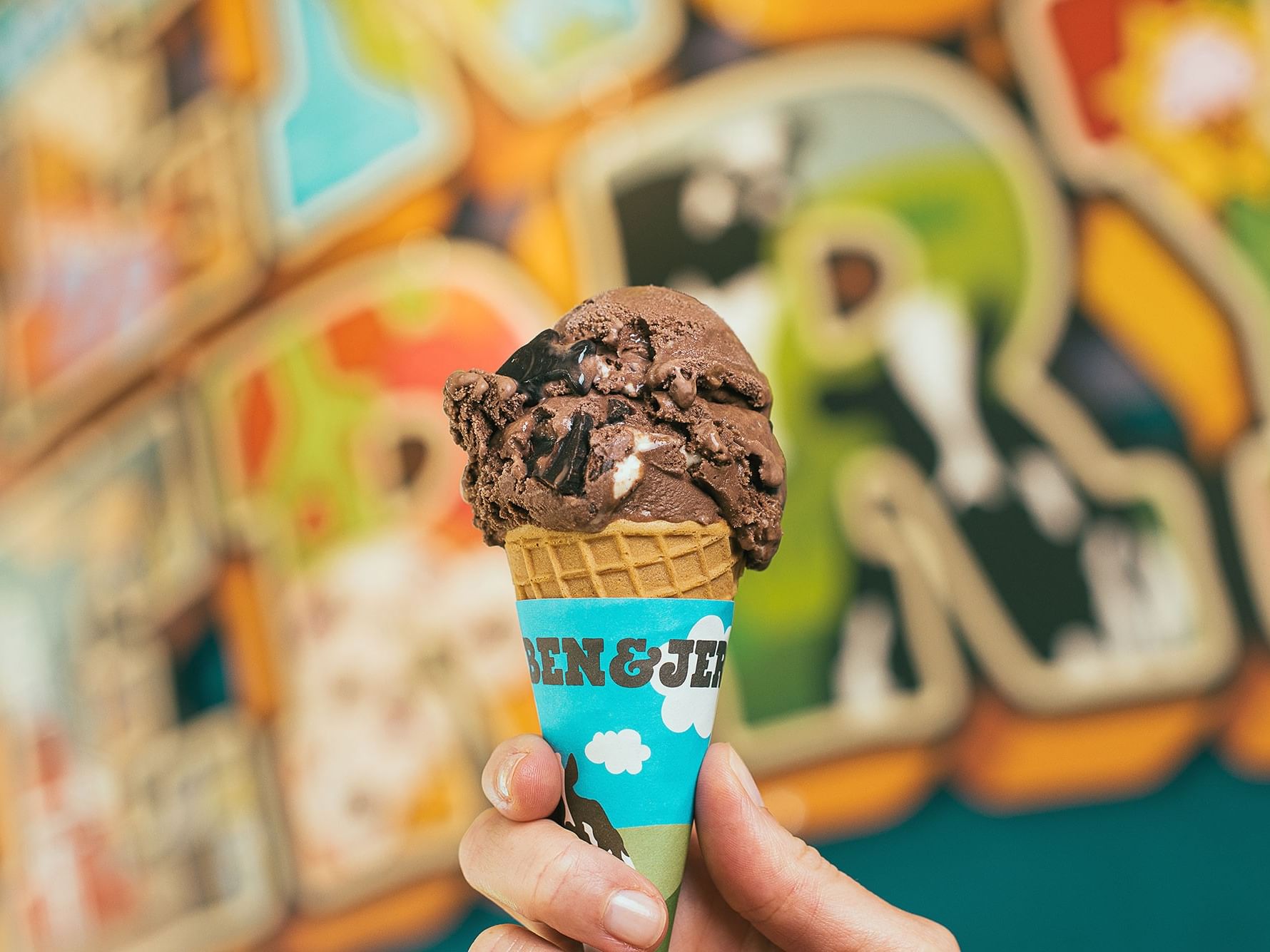 Hand holding a Ben & Jerry's ice cream cone with chocolate and cookie chunks, against a colorful mural background.