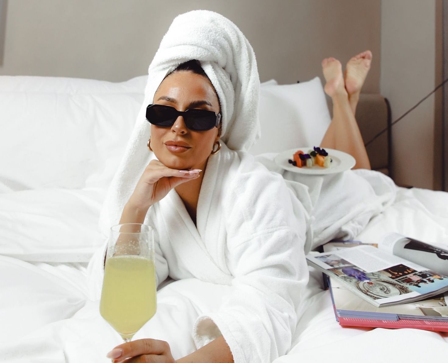 Lady wearing sunglasses and a bathrobe, posing for a picture while enjoying juice on the bed at The Londoner