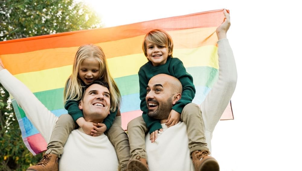 Two smiling men with children sitting on their shoulders holding a rainbow pride flag.