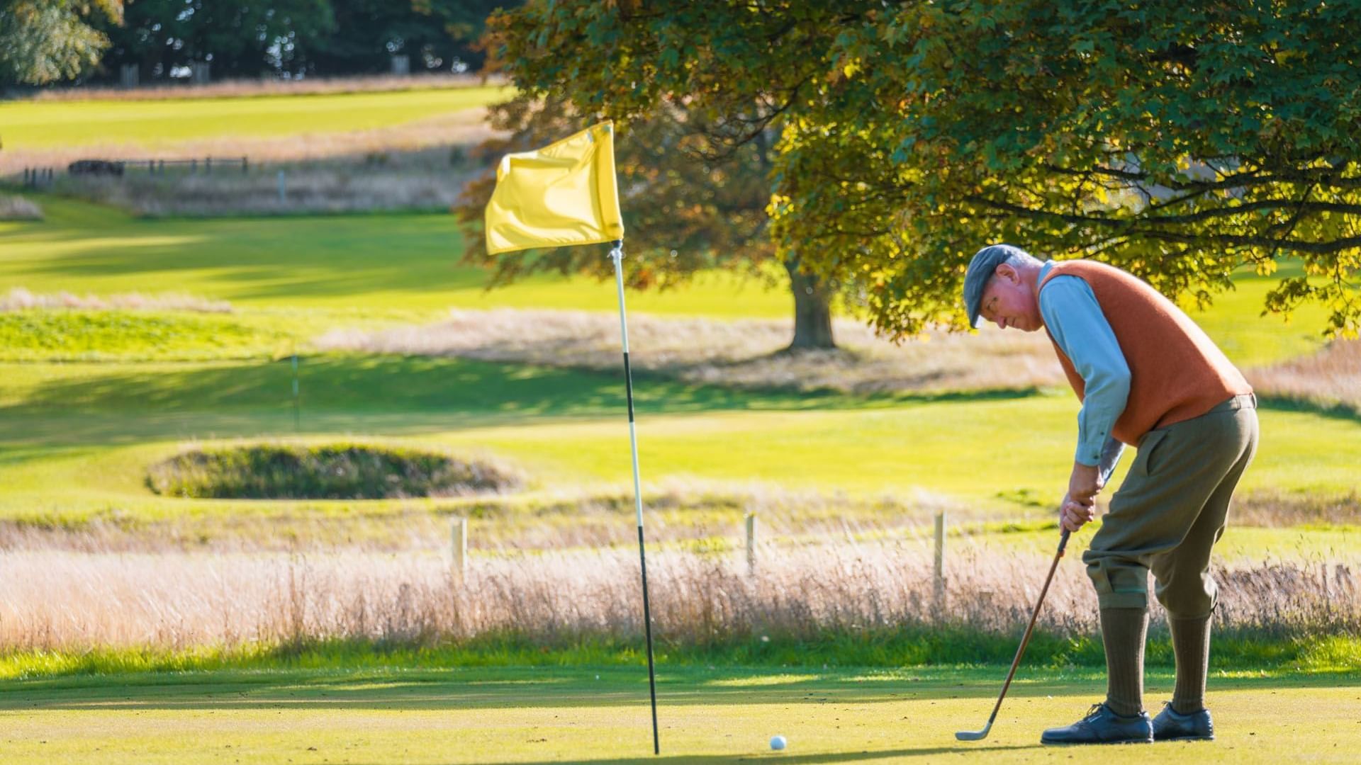 Golfer in mid-swing on a sunny golf course aiming near a hole with a yellow flag near Hotel St Andrews Golf