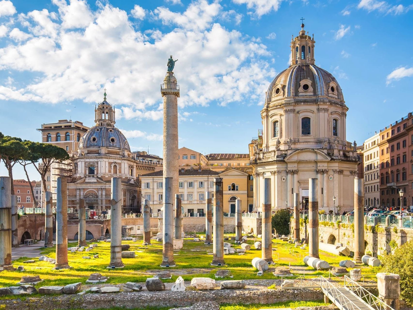 Trajan's Forum ruins with columns, twin domed churches, and a clear blue sky on a sunny day near The Guardian Hotel