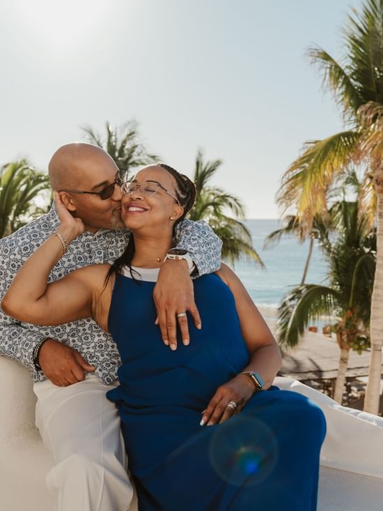 A man kisses a smiling woman on a sunny balcony, overlooking the beach at Marquis Los Cabos Resort surrounded by palms