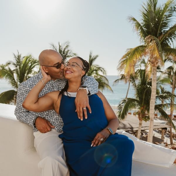 A man kisses a smiling woman on a sunny balcony, overlooking the beach at Marquis Los Cabos Resort surrounded by palms