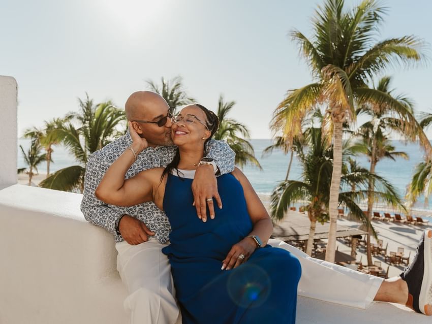 A man kisses a smiling woman on a sunny balcony, overlooking the beach at Marquis Los Cabos Resort surrounded by palms