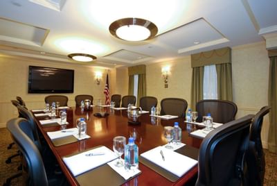 Board Room with long table, chairs, papers, bottles, flag, TV, and lamps on the ceiling.