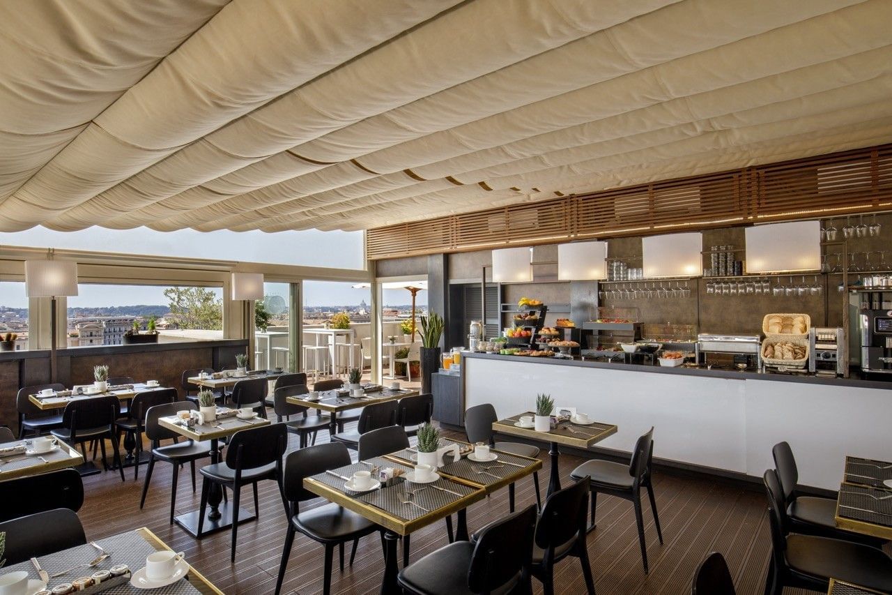 Modern rooftop dining area with neatly set tables, black chairs, and a breakfast buffet counter, offering panoramic city views through large windows.